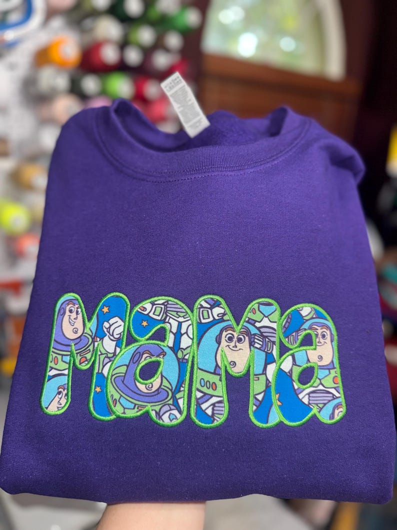 Puede incluir: Una sudadera morada de cuello redondo con la palabra "mama" bordada en una fuente de color verde brillante. El interior de cada letra est&aacute; relleno con un patr&oacute;n gr&aacute;fico colorido que representa al personaje Buzz Lightyear de Toy Story.