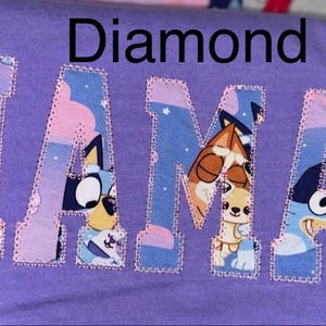 May include: A purple t-shirt featuring the word MAMA appliqued in a block font. The letters are filled with a colorful cartoon pattern of blue and orange dogs against a starry sky background, outlined with a decorative pink stitched border.