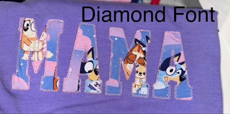 May include: A purple t-shirt featuring the word MAMA appliqued in a block font. The letters are filled with a colorful cartoon pattern of blue and orange dogs against a starry sky background, outlined with a decorative pink stitched border.
