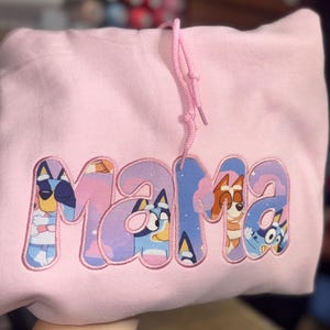 May include: A light pink hooded sweatshirt featuring the word MAMA embroidered in large letters. The letters are filled with a colorful cartoon pattern depicting blue and orange dog characters against a background of light purple clouds and stars.
