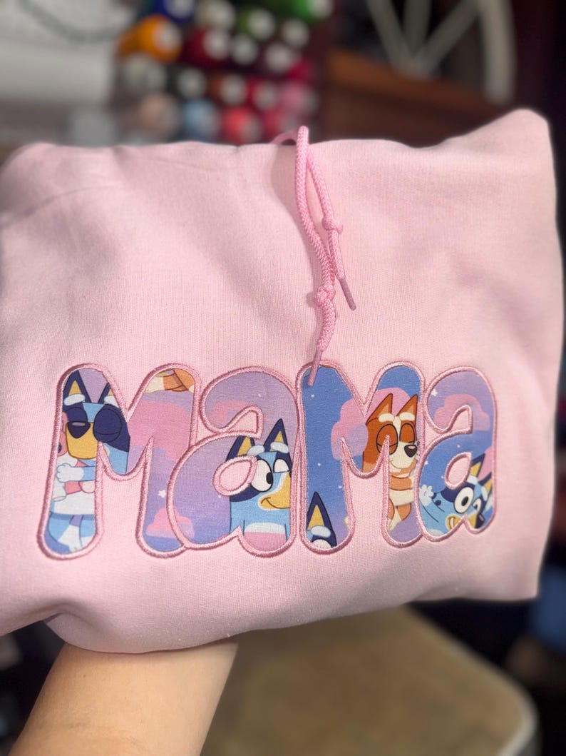 May include: A light pink hooded sweatshirt featuring the word MAMA embroidered in large letters. The letters are filled with a colorful cartoon pattern depicting blue and orange dog characters against a background of light purple clouds and stars.