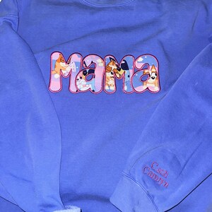 May include: A periwinkle blue crewneck sweatshirt featuring the word MAMA in large, pink-outlined letters filled with a cartoon dog pattern. The sleeve is embroidered with the names Cash and Camryn in pink thread. A Comfort Colors brand label is visible.