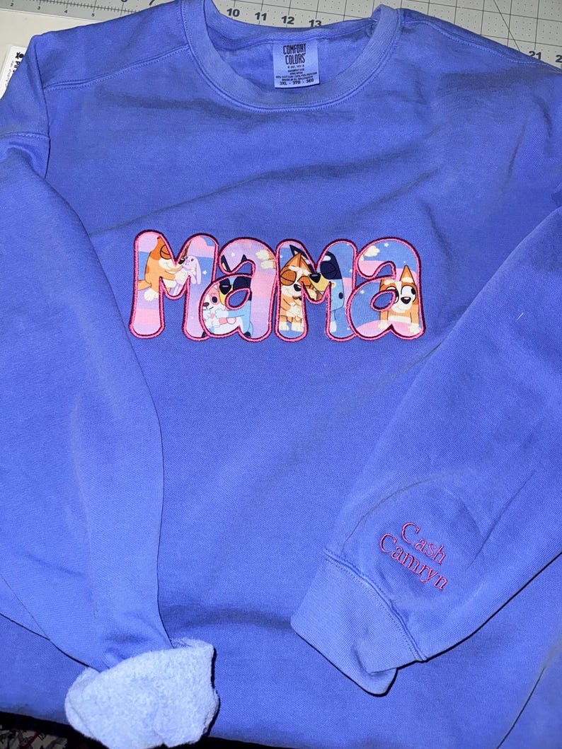 May include: A periwinkle blue crewneck sweatshirt featuring the word MAMA in large, pink-outlined letters filled with a cartoon dog pattern. The sleeve is embroidered with the names Cash and Camryn in pink thread. A Comfort Colors brand label is visible.