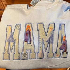 May include: A light blue crewneck sweatshirt featuring the word MAMA in large block letters. The letters are filled with a patterned fabric depicting characters from the movie Frozen, including Anna, Elsa, and Olaf, outlined in blue embroidery.