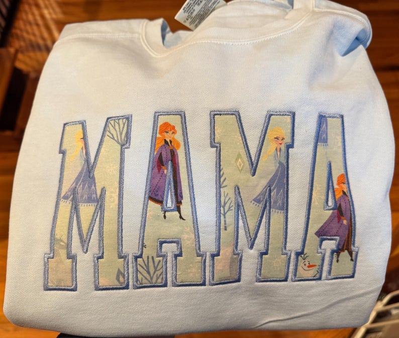 May include: A light blue crewneck sweatshirt featuring the word MAMA in large block letters. The letters are filled with a patterned fabric depicting characters from the movie Frozen, including Anna, Elsa, and Olaf, outlined in blue embroidery.