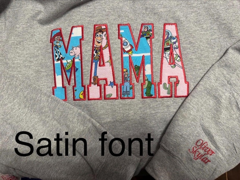 May include: A heather gray sweatshirt featuring the word MAMA in large block letters with a red stitched outline. The letters are filled with a pattern of Toy Story characters and motifs. The sleeve includes the embroidered name Oliver Skylar.