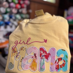 Op de afbeelding: Een lichtgeel T-shirt met de geborduurde tekst "girl" in roze cursief boven het woord "mama" in grote blokletters. Elke letter van "mama" is gevuld met een ander kleurrijk patroon met afbeeldingen van verschillende animatiefiguren.
