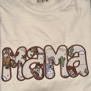 Puede incluir: Una camiseta de color crema con la palabra MAMA en letras grandes con contorno marr&oacute;n. Dentro de las letras hay ilustraciones coloridas de varios personajes de dibujos animados, incluyendo un vaquero, una vaquera, un caballo y una pastora.