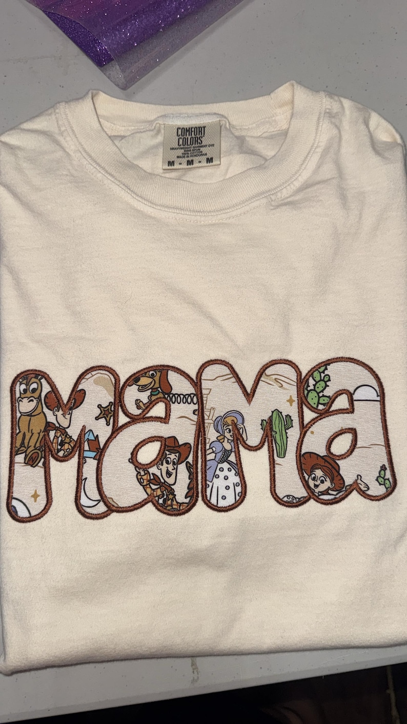 Puede incluir: Una camiseta de color crema con la palabra MAMA en letras grandes con contorno marr&oacute;n. Dentro de las letras hay ilustraciones coloridas de varios personajes de dibujos animados, incluyendo un vaquero, una vaquera, un caballo y una pastora.