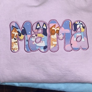 May include: A light purple sweatshirt featuring the word MAMA embroidered in large, bubble-style letters. The interior of each letter contains a colorful graphic print of cartoon dog characters from the show Bluey against a starry night sky.