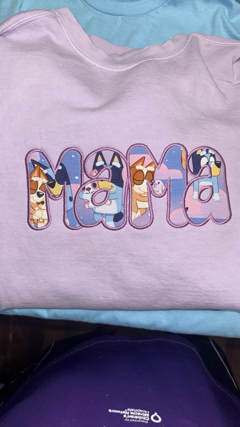 May include: A light purple sweatshirt featuring the word MAMA embroidered in large, bubble-style letters. The interior of each letter contains a colorful graphic print of cartoon dog characters from the show Bluey against a starry night sky.
