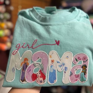 Op de afbeelding: Een lichtblauw t-shirt met de geborduurde tekst "girl mama" in roze cursief boven het woord "MAMA" in grote blokletters. Elke letter van "MAMA" bevat een andere gedrukte afbeelding van een vrouwelijk geanimeerd personage.