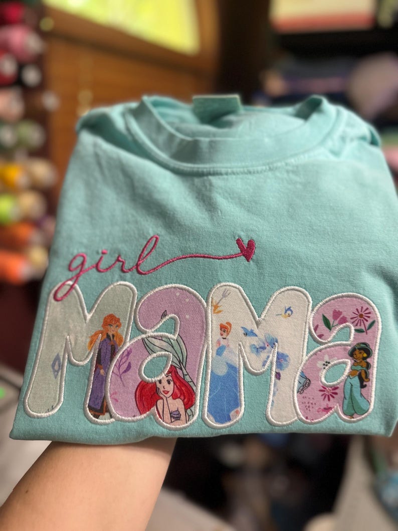 Op de afbeelding: Een lichtblauw t-shirt met de geborduurde tekst "girl mama" in roze cursief boven het woord "MAMA" in grote blokletters. Elke letter van "MAMA" bevat een andere gedrukte afbeelding van een vrouwelijk geanimeerd personage.