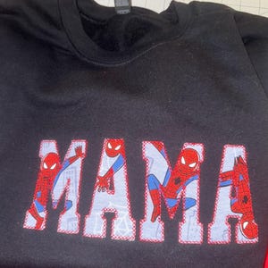 K&ouml;nnte beinhalten: Ein schwarzes Sweatshirt mit Rundhalsausschnitt und dem Wort MAMA in gro&szlig;en Blockbuchstaben. Die Buchstaben sind mit einem hellblauen Stoffmuster gef&uuml;llt, das die Figur Spider-Man in verschiedenen Posen zeigt, umrandet mit roter Naht.