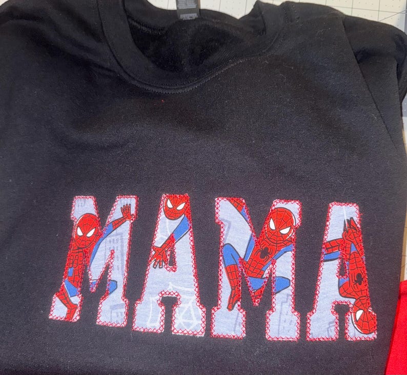 K&ouml;nnte beinhalten: Ein schwarzes Sweatshirt mit Rundhalsausschnitt und dem Wort MAMA in gro&szlig;en Blockbuchstaben. Die Buchstaben sind mit einem hellblauen Stoffmuster gef&uuml;llt, das die Figur Spider-Man in verschiedenen Posen zeigt, umrandet mit roter Naht.