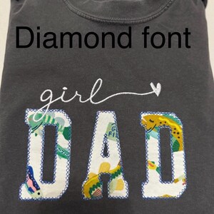 K&ouml;nnte beinhalten: Ein anthrazitfarbenes T-Shirt mit dem Text Diamond font oben. Darunter ist das Wort girl in wei&szlig;er Schreibschrift mit einem Herz gestickt. Das Wort DAD ist in gro&szlig;en Blockbuchstaben mit einem bunten Fischmuster und blauen N&auml;hten appliziert.