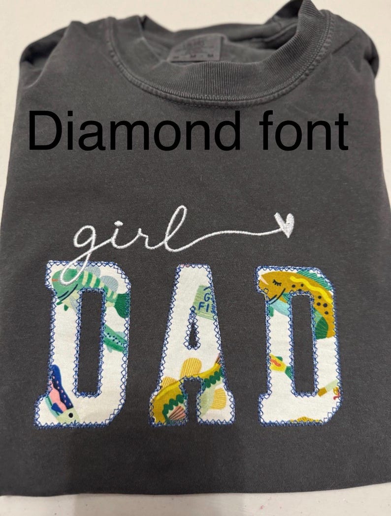 K&ouml;nnte beinhalten: Ein anthrazitfarbenes T-Shirt mit dem Text Diamond font oben. Darunter ist das Wort girl in wei&szlig;er Schreibschrift mit einem Herz gestickt. Das Wort DAD ist in gro&szlig;en Blockbuchstaben mit einem bunten Fischmuster und blauen N&auml;hten appliziert.