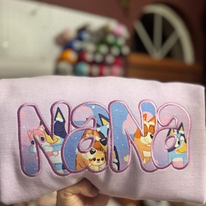 May include: A light purple sweatshirt featuring the word NANA appliqued in large, bold letters. The letters are filled with a colorful graphic print of cartoon dogs from the Bluey series against a starry night sky and have a purple stitched border.