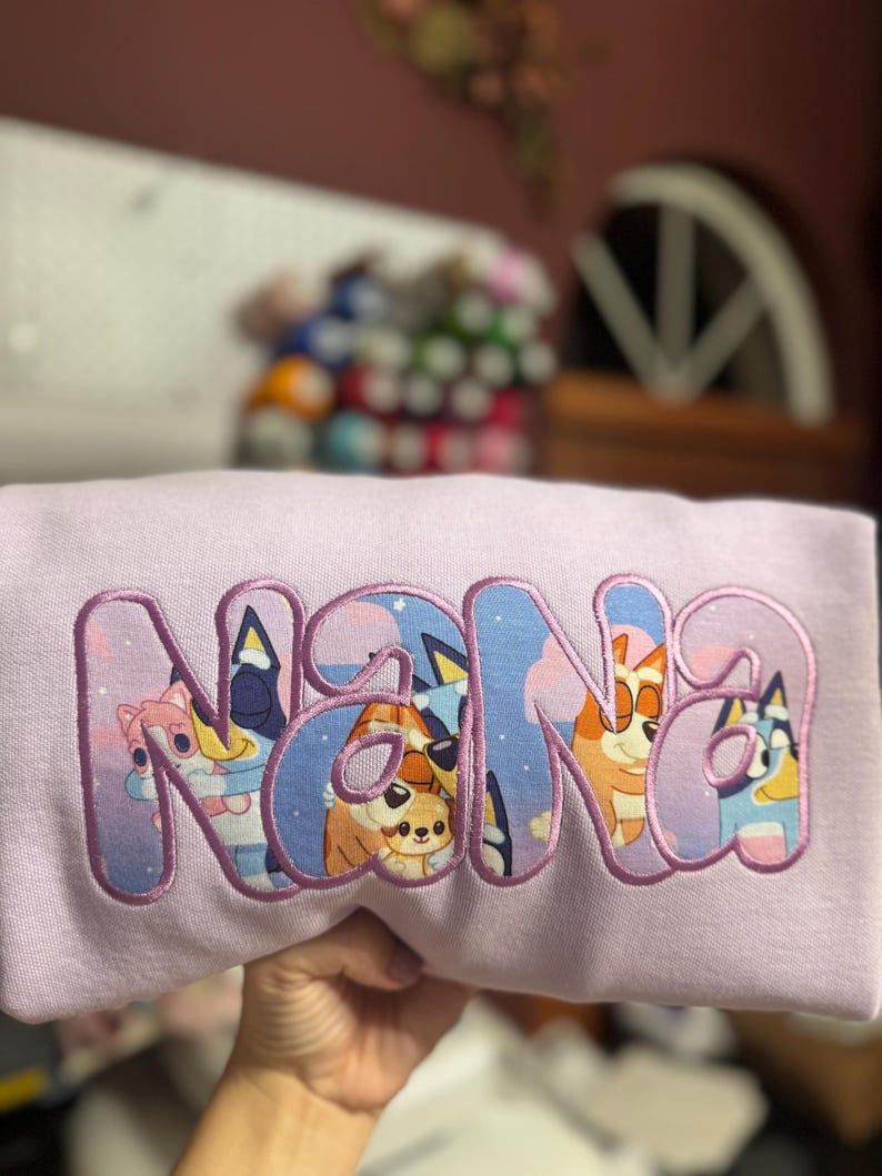 May include: A light purple sweatshirt featuring the word NANA appliqued in large, bold letters. The letters are filled with a colorful graphic print of cartoon dogs from the Bluey series against a starry night sky and have a purple stitched border.