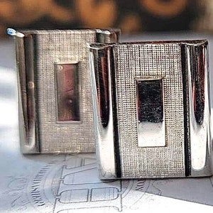 May include: A pair of silver cufflinks with a textured finish and a rectangular design. Each cufflink has a smaller rectangular inset in the center.