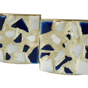 May include: A pair of square cufflinks with a gold-colored border. The face of each cufflink features a mosaic design of white, navy blue, and beige-colored pieces.