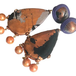 May include: A pair of handcrafted earrings featuring a copper-colored teardrop shape with black and silver accents. The earrings have a purple circular top and are adorned with dangling orange-toned beads, connected by gold-colored chains.