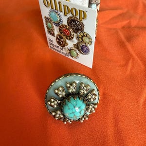 May include: A large, round ring with a turquoise stone center, surrounded by small pearls and a light blue base. The ring is set on an orange surface, with a card that reads "Ollipop" and "the pop art of personal style."