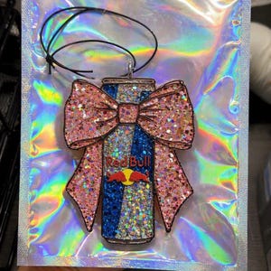 May include: A glittery Red Bull can-shaped air freshener with a pink bow. The can features the Red Bull logo and is decorated with blue and silver glitter. The bow is also covered in pink glitter. A black string is attached for hanging.
