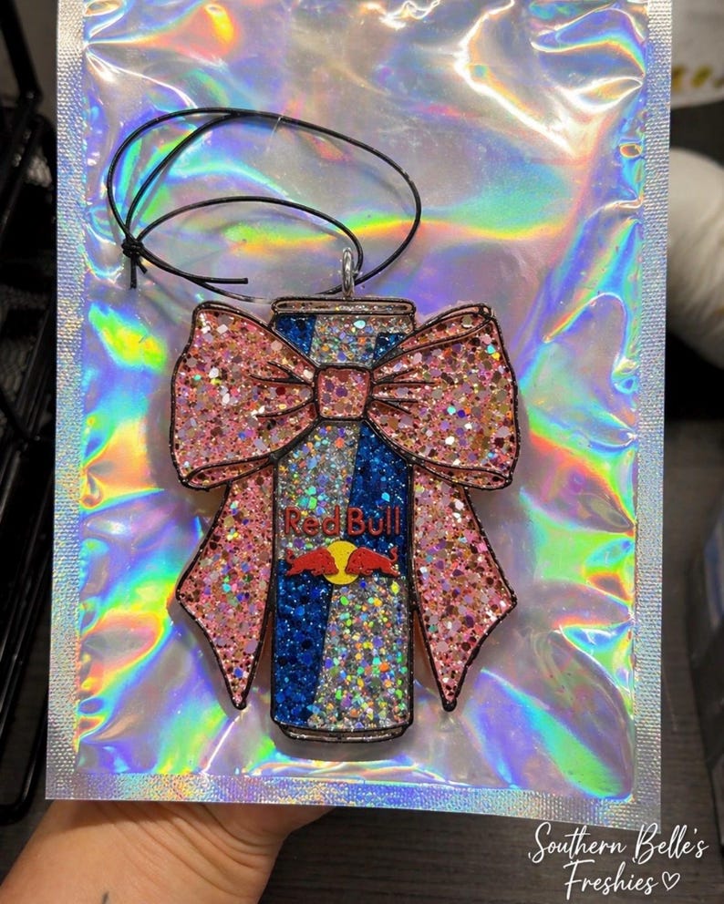 May include: A glittery Red Bull can-shaped air freshener with a pink bow. The can features the Red Bull logo and is decorated with blue and silver glitter. The bow is also covered in pink glitter. A black string is attached for hanging.