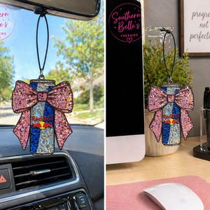 May include: A glittery air freshener shaped like a Red Bull can with a pink bow. The can has blue and silver stripes with the Red Bull logo. The air freshener is hanging from a black string. The text "Southern Belle's Freshies" is in a circle.