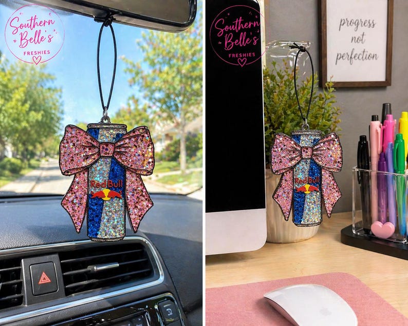 May include: A glittery air freshener shaped like a Red Bull can with a pink bow. The can has blue and silver stripes with the Red Bull logo. The air freshener is hanging from a black string. The text "Southern Belle's Freshies" is in a circle.