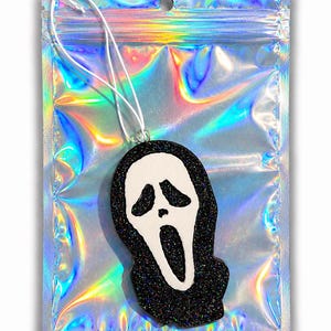 May include: A black glittery ornament shaped like a ghost face mask with a white face and a black hood. The ornament features a white string loop for hanging and is packaged inside a rectangular, iridescent, holographic plastic pouch.