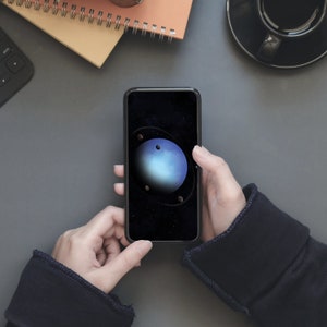 May include: A person is holding a smartphone with a dark background and a blue planet with rings around it. The planet is surrounded by a few smaller, darker planets.