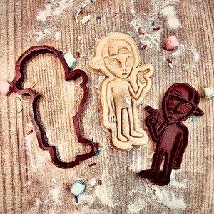 May include: Three alien-themed cookie cutters and a baked cookie on a wooden surface dusted with flour and sprinkles. The cookie cutters are in the shape of an alien, a ghost, and an alien wearing a hat. The baked cookie is in the shape of an alien.