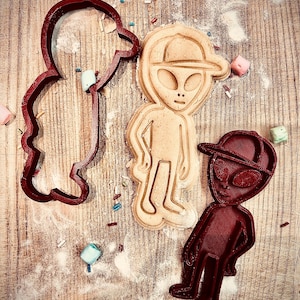 May include: Three alien-shaped cookie cutters in dark red, alongside a baked cookie in the shape of an alien wearing a cap. The scene is set on a wooden surface dusted with flour and decorated with sprinkles and marshmallows.