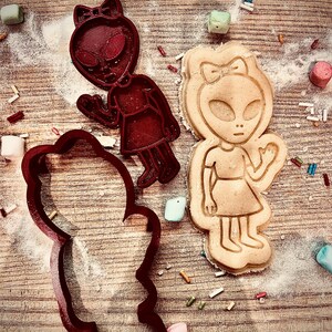 May include: Three red alien-shaped cookie cutters and a baked cookie in the shape of an alien girl. The cutters and cookie are on a wooden surface with sprinkles and marshmallows scattered around.