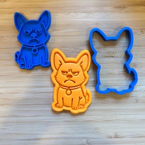 Puede incluir: Tres cortadores de galletas con forma de perro. Dos son sólidos, uno azul y otro naranja, con una expresión de mal humor. El tercero es un contorno azul de la misma forma de perro. Todo sobre una superficie de madera.