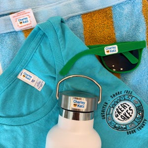 May include: A collection of summer items, including a turquoise t-shirt, green sunglasses, and a white water bottle. Each item has a label that says "Charley Katz". A blue and yellow striped towel is in the background.
