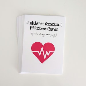 Puede incluir: Una pila de tarjetas blancas. La tarjeta superior tiene el texto "Healthcare Assistant Milestone Cards (you're doing amazing)" en negro, con un corazón rojo y un gráfico de latido blanco. Tarjetas sobre una superficie blanca.