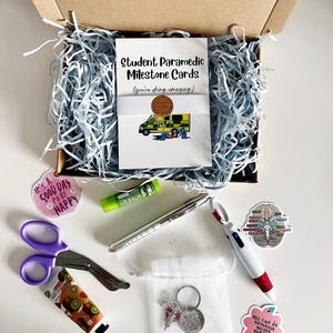 Op de afbeelding: Een geschenkdoos met een kaarten set "Student Paramedic Milestone Cards", een paarse schaar, een pen, lippenbalsem, een sleutelhanger en stickers. De doos is gevuld met blauw crêpepapier.