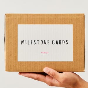 K&ouml;nnte beinhalten: Ein brauner Pappkarton mit einem wei&szlig;en Etikett mit der Aufschrift "MILESTONE CARDS" in schwarzer Schrift. Die Worte "MADE BY" und ein kleinerer Text stehen darunter. Die Box wird von einer Hand gehalten.