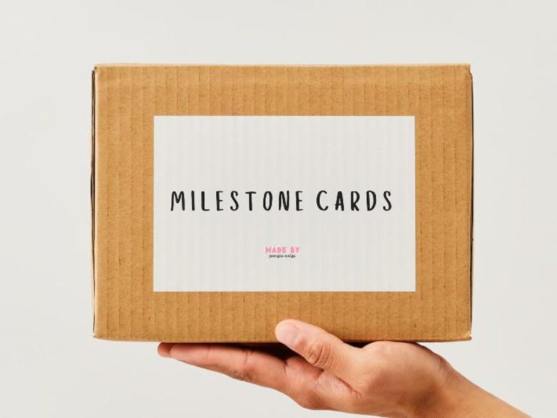 K&ouml;nnte beinhalten: Ein brauner Pappkarton mit einem wei&szlig;en Etikett mit der Aufschrift "MILESTONE CARDS" in schwarzer Schrift. Die Worte "MADE BY" und ein kleinerer Text stehen darunter. Die Box wird von einer Hand gehalten.