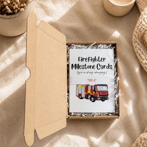 May include: A brown cardboard box containing a stack of white cards titled Firefighter Milestone Cards (you're doing amazing) with a graphic of a red and yellow fire engine. A white paper band with a circular logo wraps around the centre of the cards.