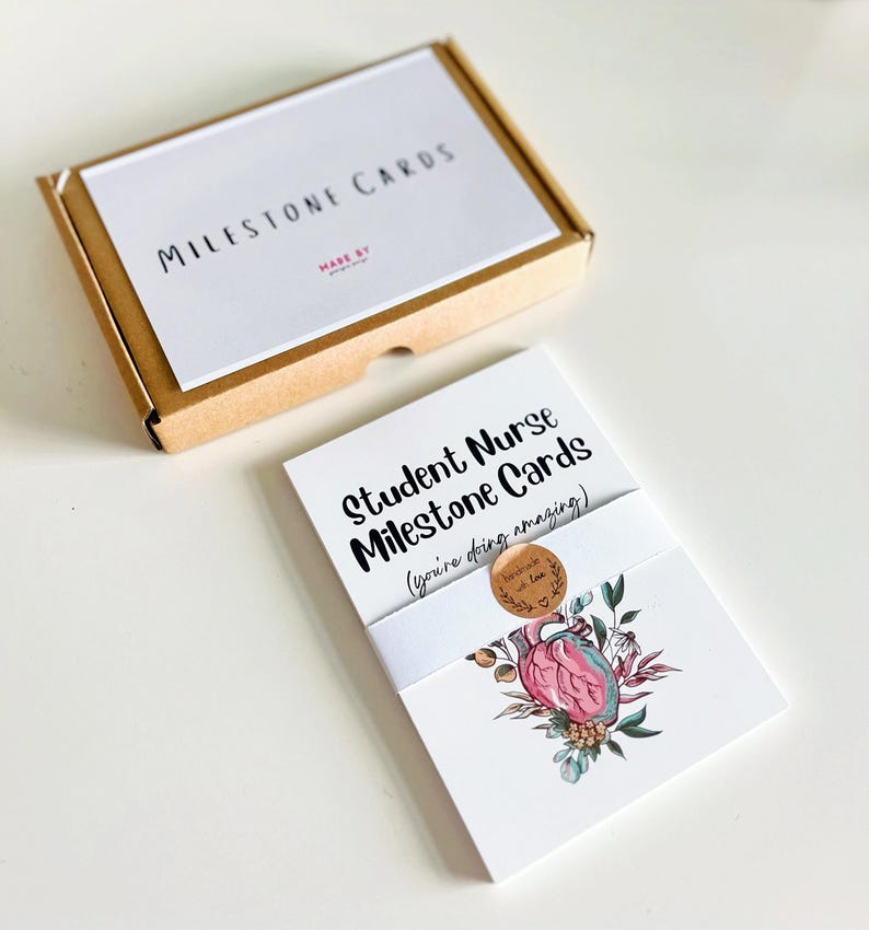 Peut inclure: Un ensemble de cartes Milestone pour &eacute;tudiants infirmiers, blanches, avec un motif de c&oelig;ur floral. Les cartes sont envelopp&eacute;es d'une bande blanche et d'un petit autocollant. Une bo&icirc;te s&eacute;par&eacute;e avec le texte "Milestone Cards" est &eacute;galement visible.