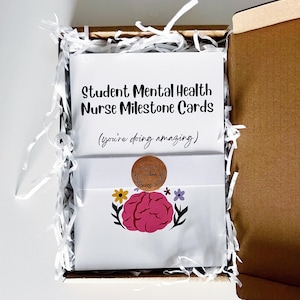 K&ouml;nnte beinhalten: Eine offene Geschenkbox aus Pappe mit wei&szlig;en Karten. Auf den Karten steht "Student Mental Health Nurse Milestone Cards" und "(you're doing amazing)". Sie sind mit einem rosa Gehirn und Blumen sowie einem Aufkleber "handmade with love" verziert.