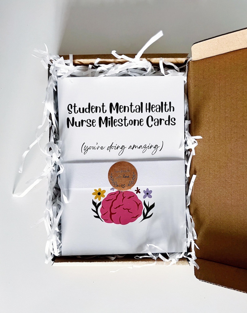 K&ouml;nnte beinhalten: Eine offene Geschenkbox aus Pappe mit wei&szlig;en Karten. Auf den Karten steht "Student Mental Health Nurse Milestone Cards" und "(you're doing amazing)". Sie sind mit einem rosa Gehirn und Blumen sowie einem Aufkleber "handmade with love" verziert.