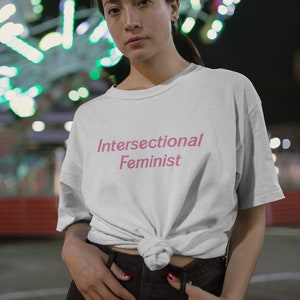 Puede incluir: Una camiseta blanca con el texto "Intersectional Feminist" impreso en rosa en la parte delantera. La camiseta está anudada a la cintura y la persona que la lleva está de pie frente a un fondo borroso de luces.