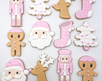 Puede incluir: Una colección de galletas de Navidad decoradas en varias formas, incluyendo un cascanueces, un árbol de Navidad, un reno, un calcetín, Papá Noel y un hombre de jengibre. Las galletas son principalmente rosas y blancas, con acentos marrones y beige. Están dispuestas sobre una superficie blanca.