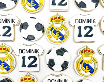 Könnte beinhalten: Ein Set mit 12 dekorierten Zuckerkeksen mit einem Fußball, einem Fußballtrikot mit dem Namen "Dominik" und der Nummer "12" sowie dem Wappen von Real Madrid. Die Kekse sind weiß mit schwarzen, blauen, goldenen und roten Akzenten.