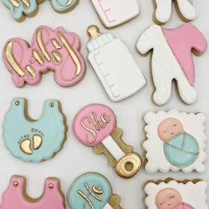 K&ouml;nnte beinhalten: Ein Set aus zehn dekorierten Zuckerkeksen mit Baby-Motiv. Die Kekse sind mit rosa, blauem und goldenem Zuckerguss verziert. Die Kekse zeigen die W&ouml;rter "Baby", "Sie", "Er" und Bilder von Babyflaschen, Stramplern und Rasseln.